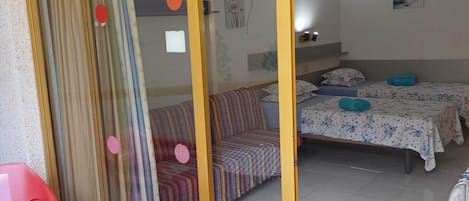 A room with a sliding glass door, a bed with a floral bedspread, a sofa with a striped blanket, and a small table with a checkered tablecloth.