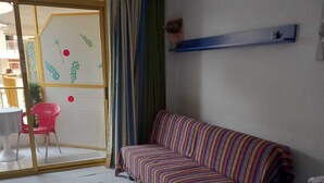 Una habitación con un sofá a rayas, una cama con colcha estampada, una almohada azul y un ventanal corredizo que da a un espacio exterior con una silla.