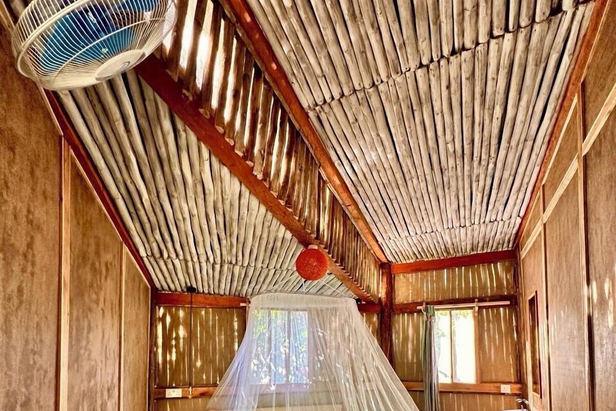 A room with a bed under a mosquito net, a wooden ceiling, and a small table with a chair.