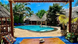 Ein Poolbereich mit Holztisch, Liegestühlen und strohgedeckten Bauwerken.