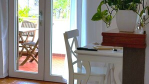 Une pièce comprenant un bureau blanc, une chaise, une plante en pot et une porte-fenêtre coulissante donnant sur un espace extérieur avec une chaise et une table en bois.