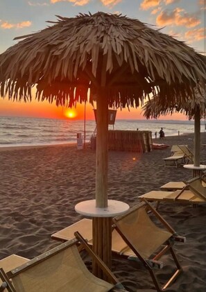 Une plage avec un parasol en chaume, des chaises longues et une vue sur le coucher de soleil.