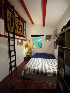 Ein Schlafzimmer mit Bett, Fenster, einer Leiter, einem Bücherregal und einem Wandbild.