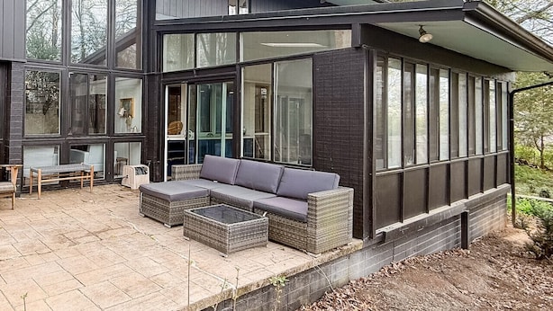 Una casa moderna con un amplio ventanal, un patio con mobiliario y una base de ladrillo.
