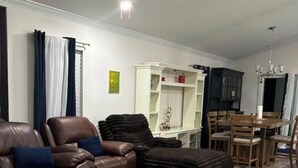 Un salon avec un canapé en cuir, une table basse en verre, un tapis à motifs, un coin repas avec des chaises en bois et une étagère blanche.