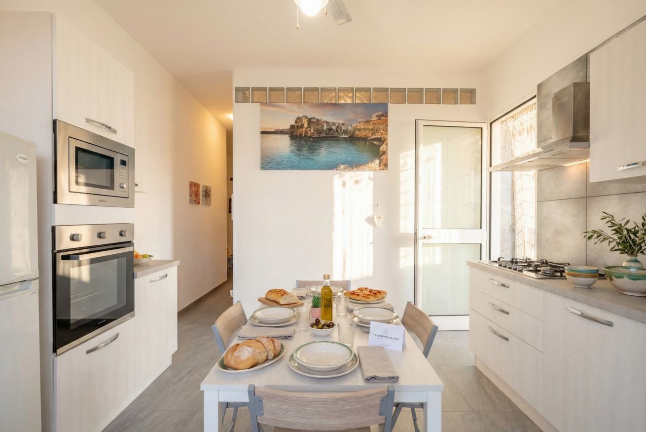 A modern kitchen with built-in appliances, a dining table set with plates, bread, and a bottle, and a coastal painting on the wall.