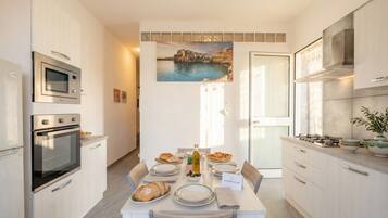 A modern kitchen with built-in appliances, a dining table set with plates, bread, and a bottle, and a coastal painting on the wall.