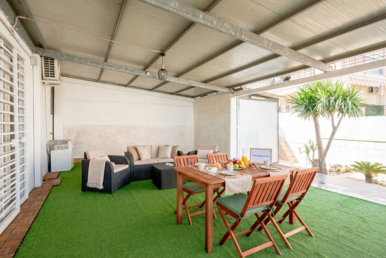 A covered outdoor area with a dining table, chairs, a sofa, and a small garden.