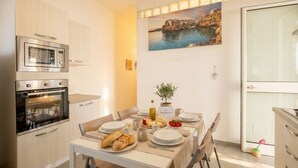 A dining area with a table set for two, a microwave, an oven, and a painting of a coastal scene.