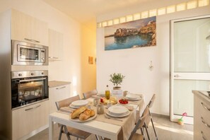 A dining area with a table set for two, a microwave, an oven, and a painting of a coastal scene. - CarmenLeo-Central Accommodation with Patio (Polignano a Mare)