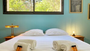 Une chambre d’hôtel comprenant un lit, deux oreillers, une table de chevet avec une lampe, une autre table de chevet avec une lampe, et une fenêtre donnant sur des arbres.