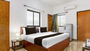 Hotellihuone, jossa on puinen sänky, pieni yöpöytä lampun kanssa, jääkaappi ja puinen vaatekaappi.