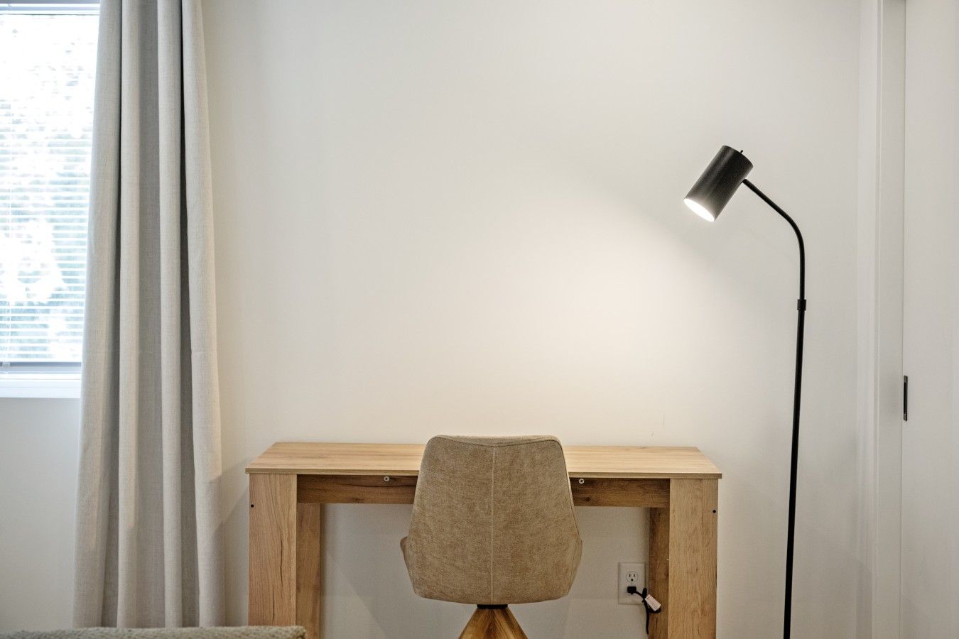 A room with a wooden desk, a chair, a floor lamp, and a window with curtains.