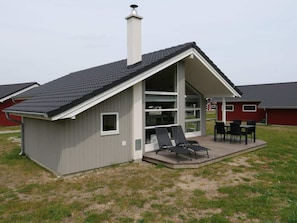 Ein kleines, eingeschossiges Gebäude mit geneigtem Dach, großen Fenstern und einer Terrasse mit Sitzgelegenheiten.
