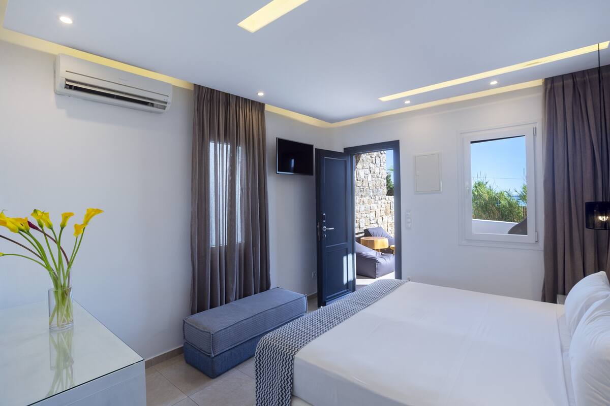 A modern hotel room with a bed, a small seating area, a TV, and an air conditioning unit.