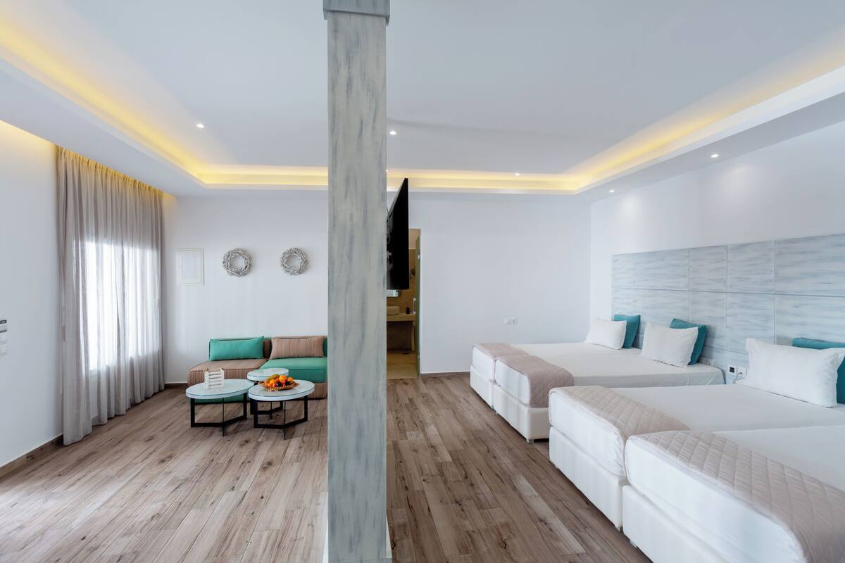 A modern hotel room with a wooden floor, a sofa with cushions, a coffee table, two beds, and a large pillar.