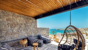 Un mur en pierre, un plafond en bois, un fauteuil suspendu en osier et un espace extérieur aménagé avec vue sur la mer.
