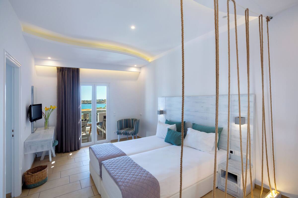 A hotel room with two beds, a desk with a TV, a chair, and a balcony with a view.