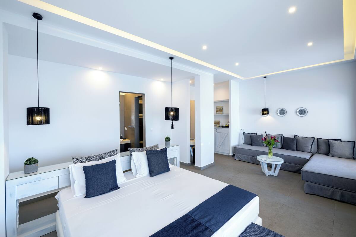 A modern hotel room with a large bed, a sofa, a small table, and a kitchen area.