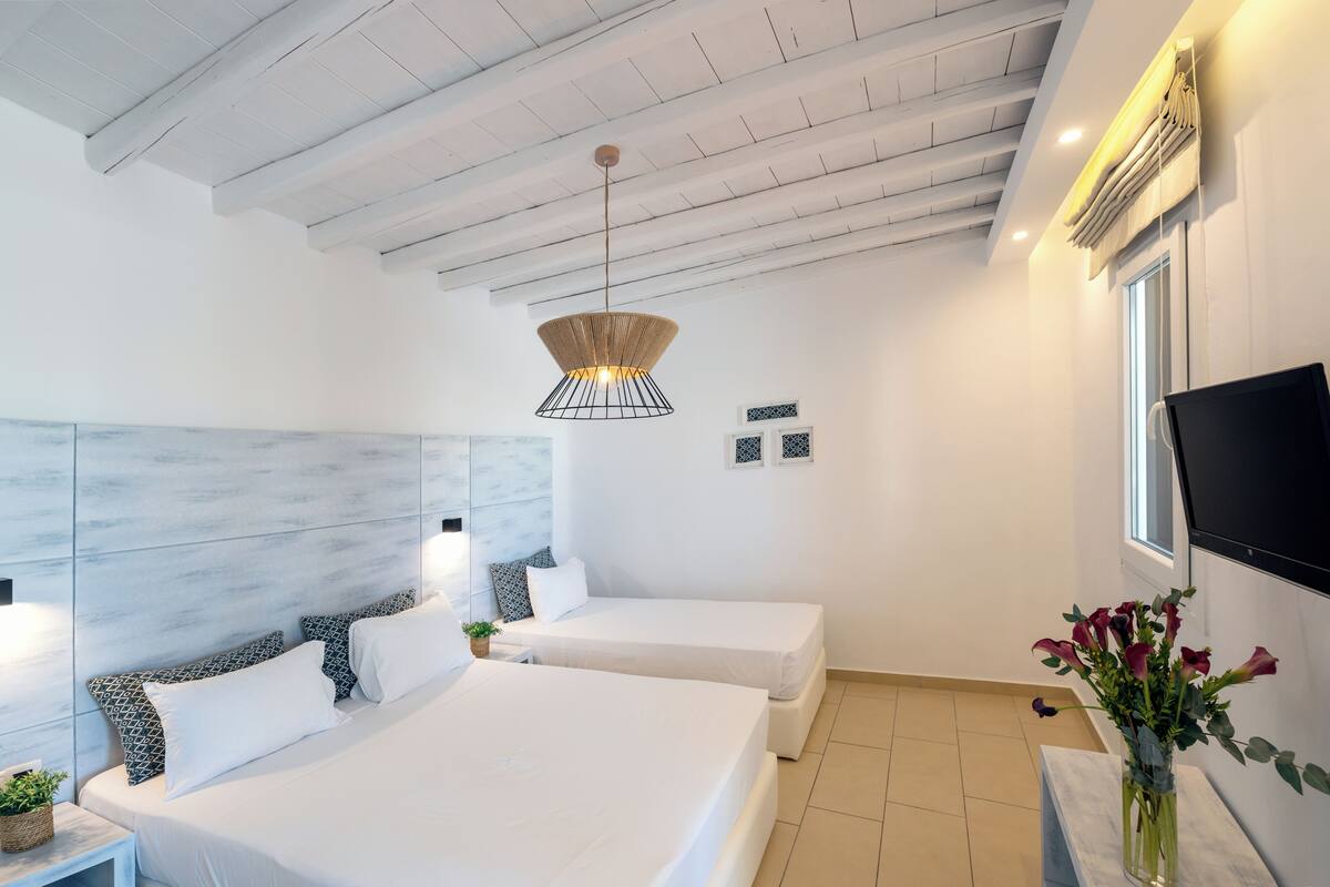 A modern hotel room with two beds, a large headboard, a hanging light fixture, a flat-screen TV, and a vase with flowers.