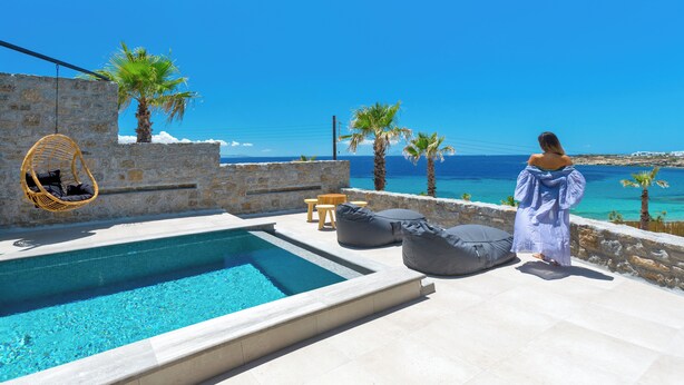 Un espace aménagé au bord de la piscine, avec un mur en pierre, un fauteuil suspendu et une personne portant une longue robe.