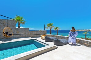 A poolside area with a stone wall, a hanging chair, and a person in a long dress. - Tropicana Hotel (Mykonos)