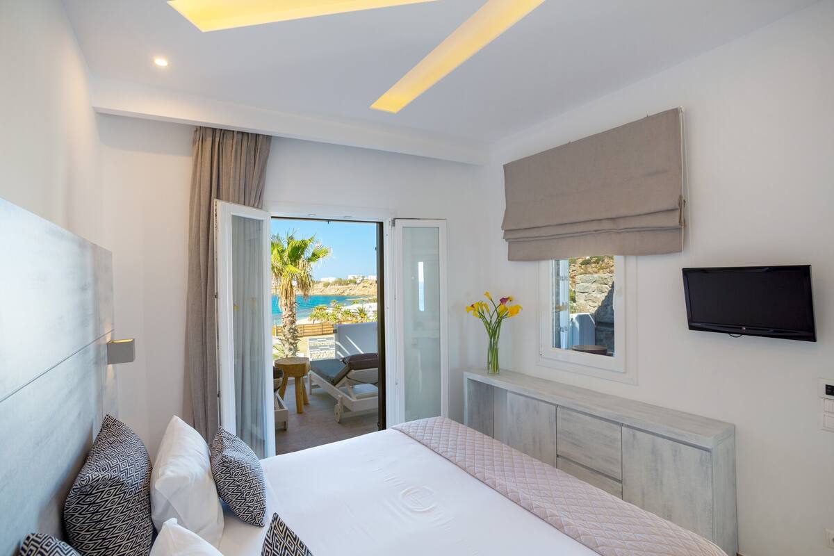 A modern hotel room with a bed, a TV, a window with a view of the sea, and a balcony with a pool.