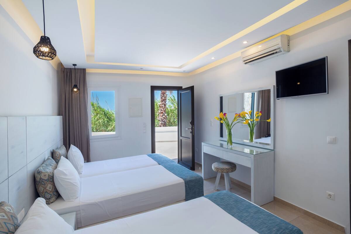 A modern hotel room with two beds, a desk, a TV, and a view of greenery outside.