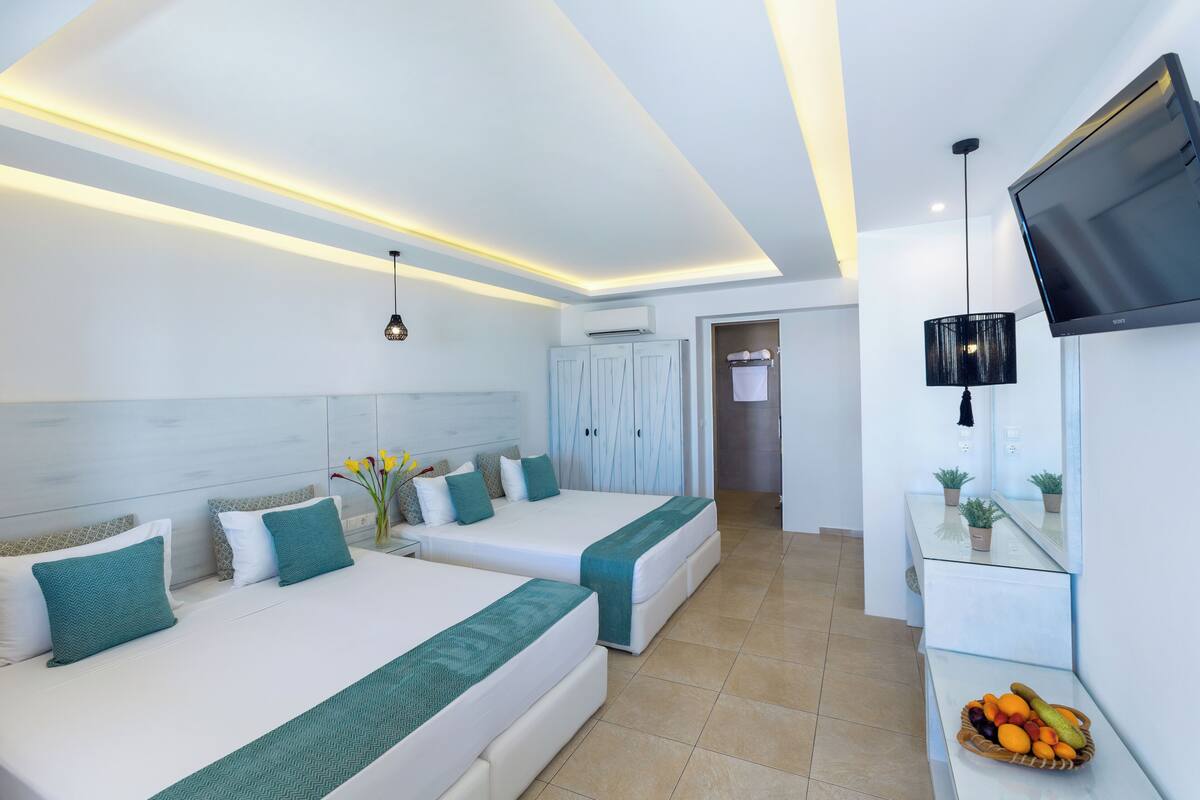 A modern hotel room with two beds, a flat-screen TV, a small table with fruit, and a bathroom visible in the background.