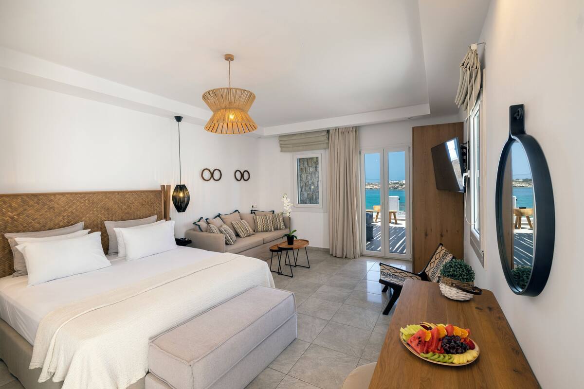 A hotel room with a large bed, a sofa, a dining table with fruit, a TV, and a view of the sea.