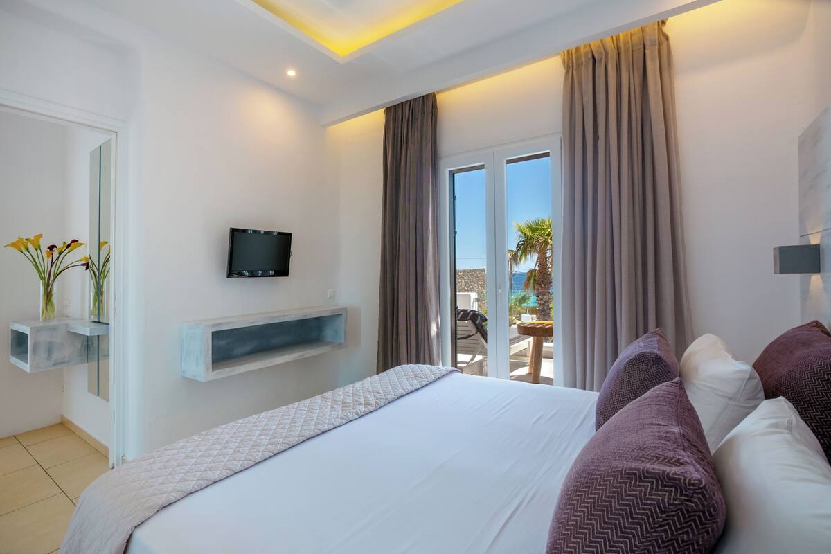 A modern hotel room with a large bed, a mounted TV, a balcony with a view, and a small table with chairs.