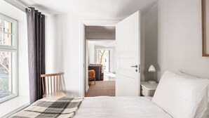 Quarto com cama, mesa de cabeceira, luminária, janela com cortinas e vista para uma sala de estar.