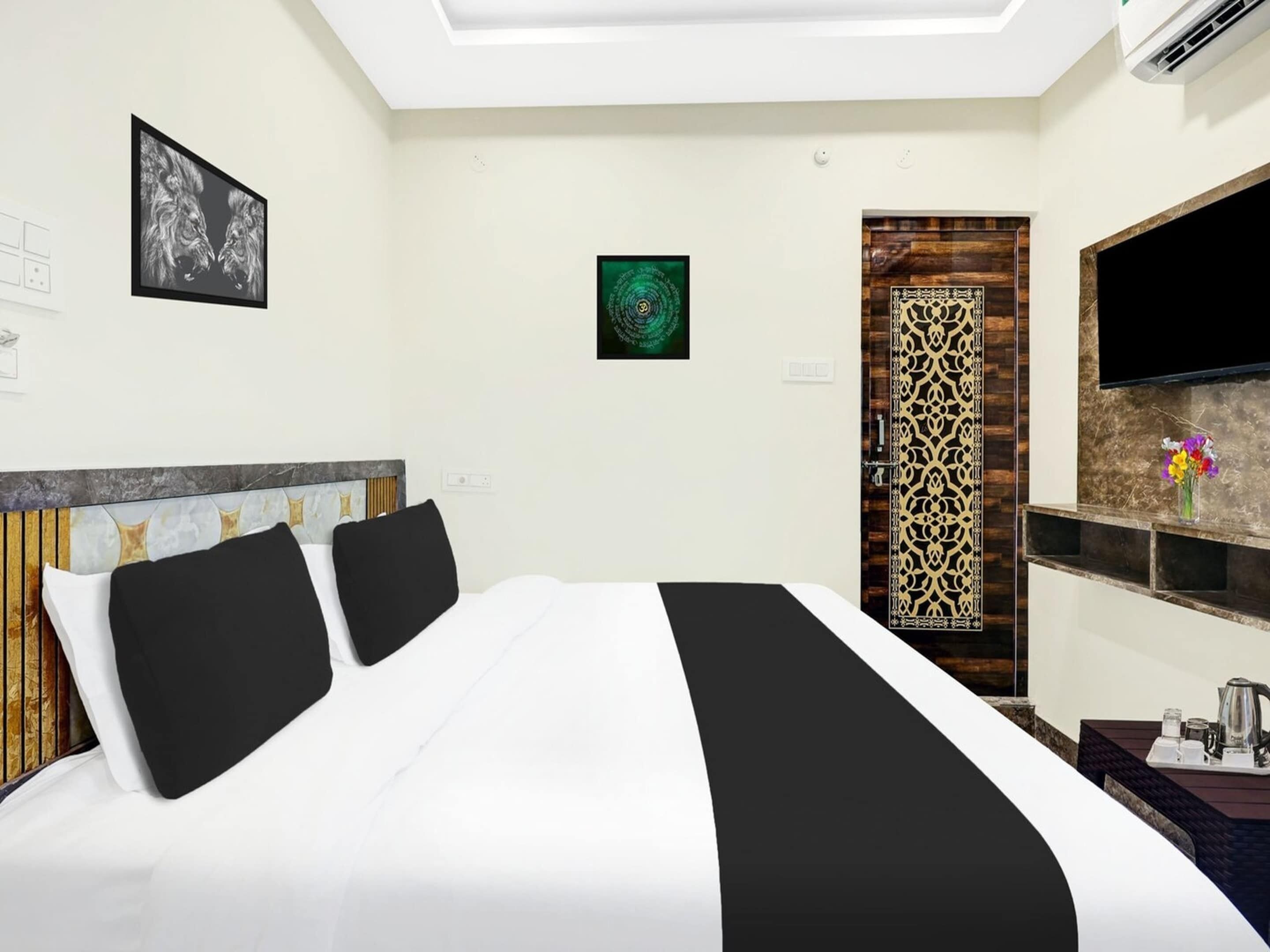 A hotel room with a bed, a TV, a decorative wall panel, and a small table with a kettle.