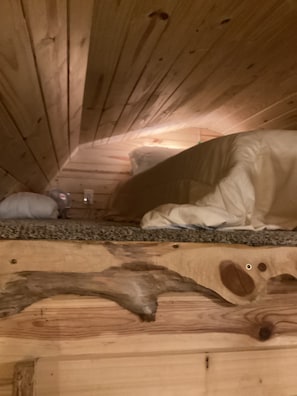 Eine Holzbalkendecke mit geneigtem Dach, einer Matratze und einem Kopfkissen auf einer strukturierten Oberfläche.