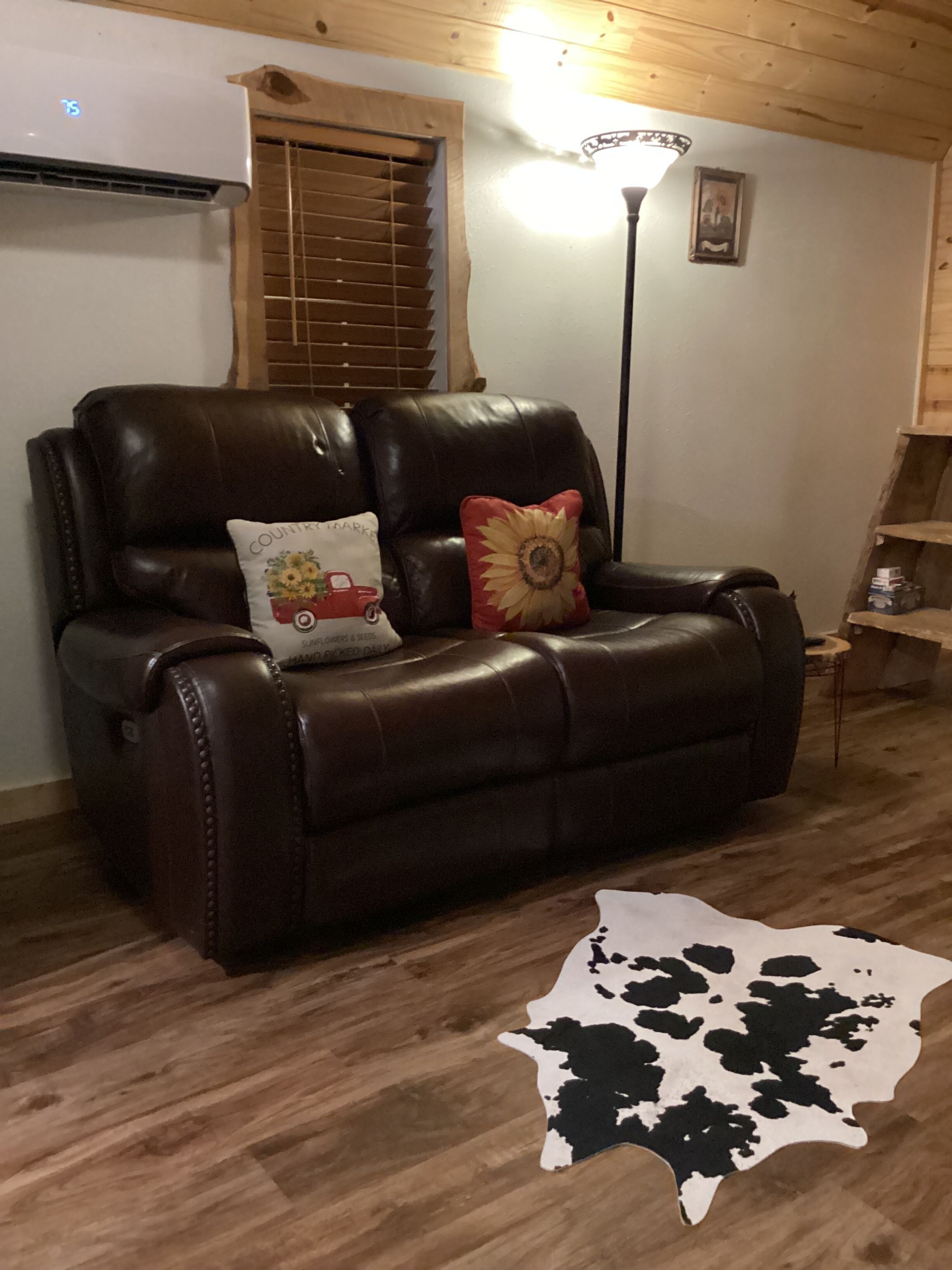 A brown leather sofa with two decorative pillows, a wooden floor, a cowhide rug, a window with blinds, and a wall-mounted air conditioner.