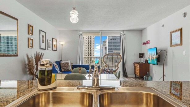 A modern kitchen with a double sink, a dining area with a blue sofa, and a view of tall buildings through large windows. - Plush Brickell 1BR | Free Parking | Resort Pool (Brickell)