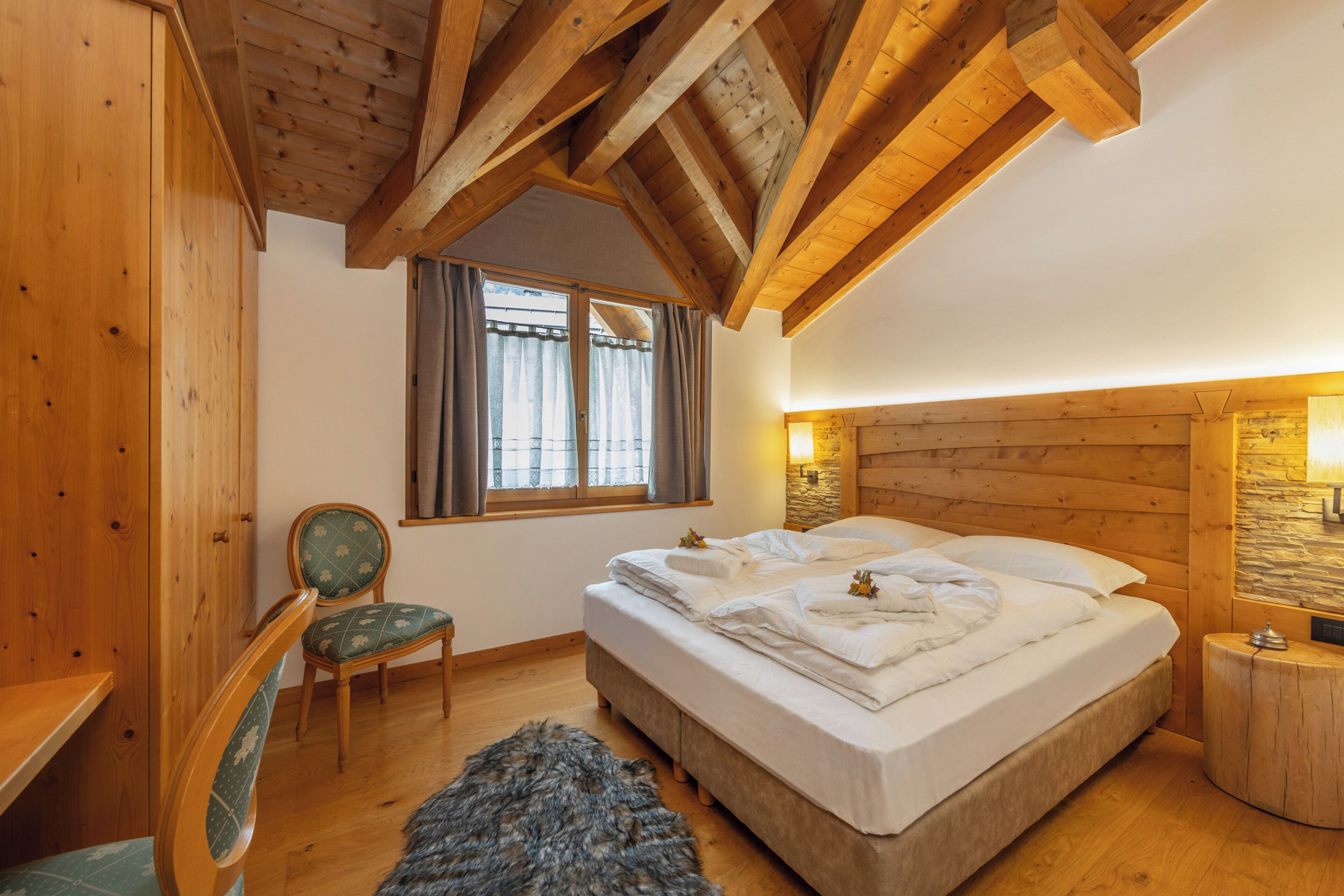 Une chambre avec un plafond en bois, un lit, une chaise, une fenêtre avec des rideaux et une table de chevet.