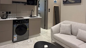 Une cuisine moderne équipée d’un four à micro-ondes, d’une machine à café et d’un lave-linge. On y trouve également un canapé et une table basse avec une télécommande et un coffre.