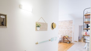 Um quarto com parede branca, uma placa de madeira com 'Bienvenue à la Maison', um chapéu, uma vassoura e uma escada com prateleiras.