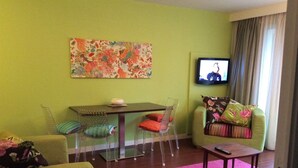 Ein Wohnzimmer mit grüner Wand, einem Tisch mit Stühlen, einer Couch, einem Fernseher und einem rosa Teppich.