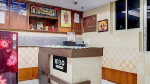 Ein Hotelrezeptionstresen mit einem Schild, das 'HOTELO & Company-Serviced' zeigt, ein Ticketschalter, ein wandmontierter Kalender und ein dekorativer Schrein.