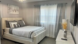 Een slaapkamer met een bed, een nachtkastje met lamp, een aan de muur bevestigde televisie en een raam met gordijnen.