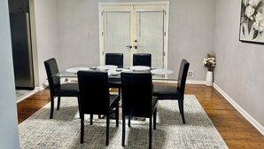 Een eetkamer met een glazen tafel, zwarte stoelen, een kroonluchter, een witte deur en een lijstwerk met bloemen aan de muur.
