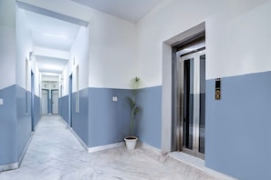 Un pasillo de hotel con piso de mármol, paredes azules y blancas, una planta en maceta y un ascensor.
