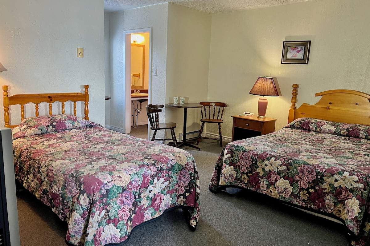 Une chambre à coucher avec deux lits en bois, un bureau, une chaise, une lampe et une salle de bain visible au fond.