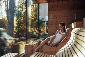 Une personne se détend dans un sauna en bois, avec vue sur les arbres à travers une grande fenêtre.