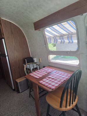 A compact kitchen with a wooden table, chairs, and a window with a view of a striped awning. - Vintage Airstream Excella in a Private Setting - Avail. May 15 through Oct. 31 (Penobscot)