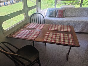 A dining area with a wooden table and four plaid placemats, a black chair, and a sofa with decorative pillows. - Vintage Airstream Excella in a Private Setting - Avail. May 15 through Oct. 31 (Penobscot)