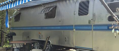 A vintage camper with a striped awning, a table, and a chair.