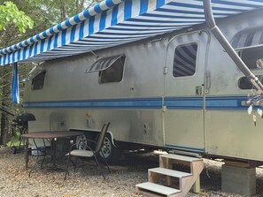 A vintage camper with a striped awning, a table, and a chair. - Vintage Airstream Excella in a Private Setting - Avail. May 15 through Oct. 31 (Penobscot)